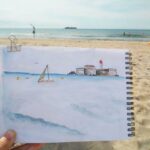 Un carnet de croquis représentant un paysage de plage est tenu devant le même paysage