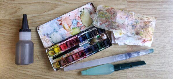Un petit pot fermé pour l'eau, une boîte d'aquarelle, un mouchoir qui fait office de chiffon et deux pinceaux à réservoir d'eau.