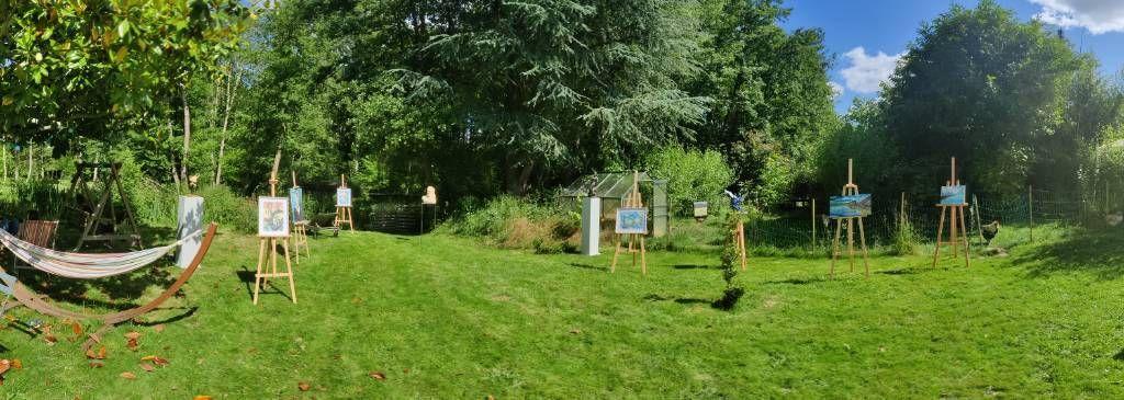 Panorama d'un jardin arboré où l'on aperçoit un hamac, une balancelle, des tableaux sur chevalets et des sculptures sur socles