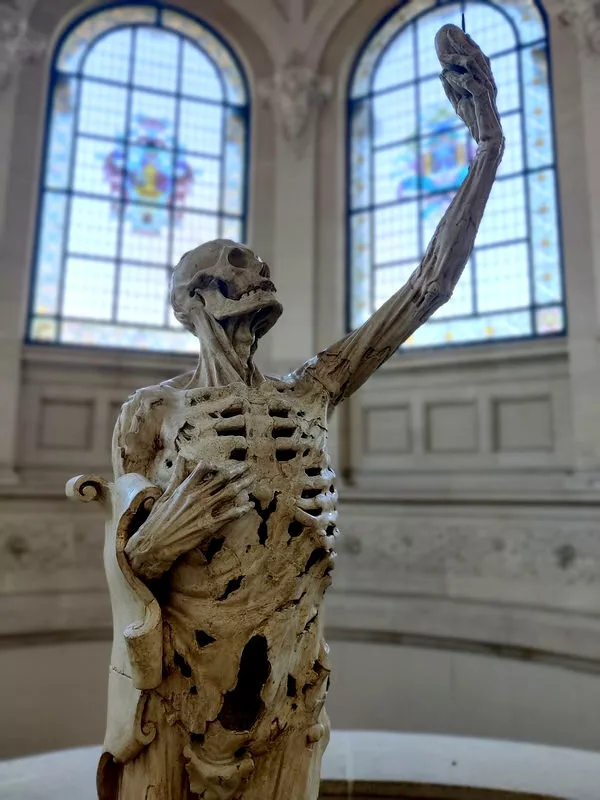 Sculpture d'un squelette en grande partie décharné regardant son cœur qu'il tient dans sa main, bras tendu vers le haut. Dans le fond, des vitraux