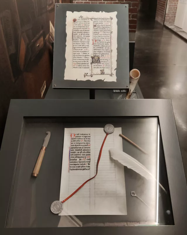 Une page de manuscrit surplombe une installation où on voit une feuille où la page est partiellement recopiée et des outils dont une plume et un calame