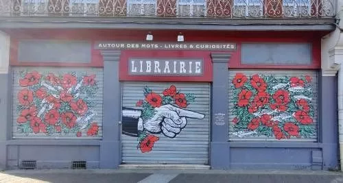 Façade d'une librairie dont les stores sont peints de fleurs rouge et d'une main pointant vers les horaires d'ouverture Au dessus du mot libraire, un texte "Autour des mots - Livres & curiosité"