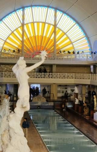 Un vitrail en arc de cercle faisant penser à un lever de soleil en arrière plan, une salle où l'on devine un petit bassin et des sculptures. Au premier plan, une sculpture d'une femme brandissant une étoile