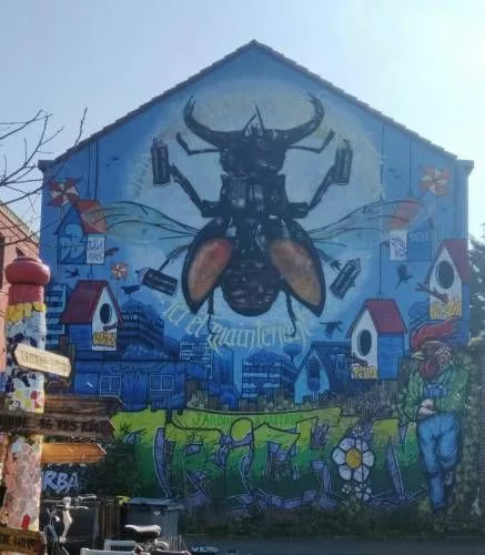 Sur toute une façade d'immeuble, une fresque avec un scarabée tenant des aérosols avec quatre de ses pattes, des nichoirs, un homme à tête de coq, des fleurs sur un fond de ville
