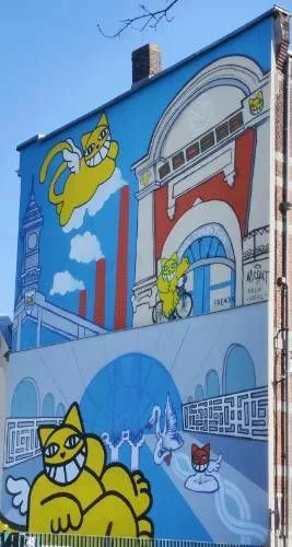 Sur toute une façade d'immeuble, une fresque colorée représentant deux saynètes avec des références à Roubaix : musée La Piscine, cheminées, La Condition Publique, etc. Un chat joyeux, jaune et ailé est présent à chaque fois