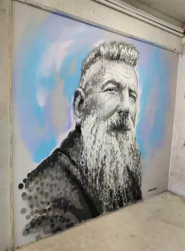 Graff occupant toute la hauteur d'un mur représentant le buste d'un homme barbu, en noir et blanc sur un fond à traces bleues, blanches et mauves "@akajimmyc"