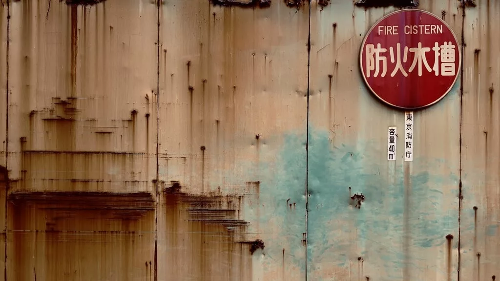 un mur rouillé avec des tons bleus et marrons ainsi qu'un panneau rouge pour annoncer un dispositif contre les incendies (en anglais et japonais).