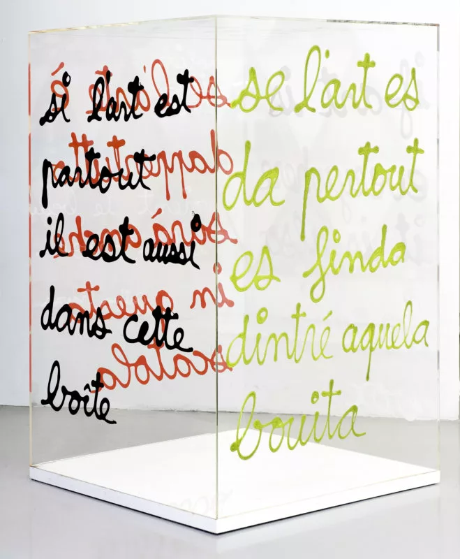 Une boîte transparent sur lequel est écrit "si l'art est partout, il est aussi dans cette boîte". La phrase est écrite sur les quatre faces, à chaque fois dans une langue et une couleur différente