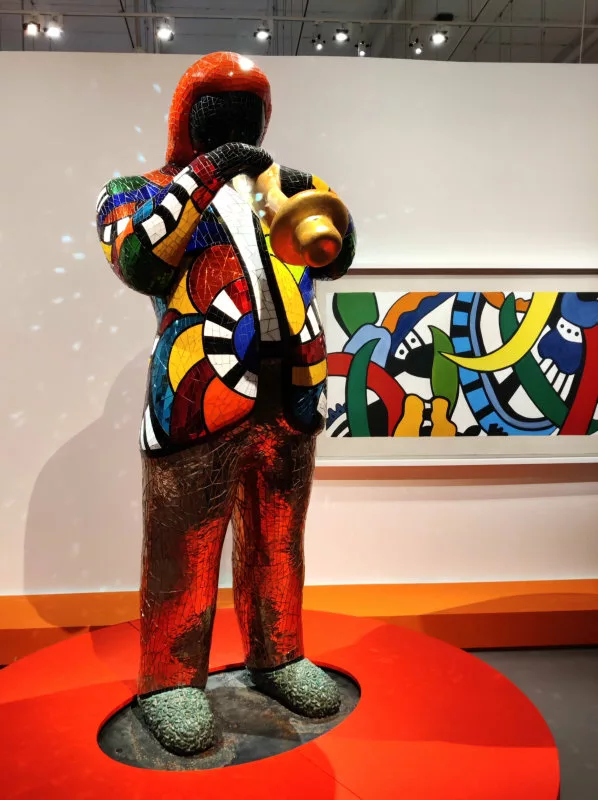 Une sculpture recouverte de mosaïque miroir colorées représentant un homme en veste en train de jouer de la trompette (et dans le fond un tableau)