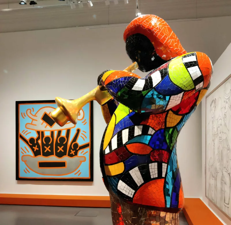 Une sculpture recouverte de mosaïque miroir colorées représentant un homme en veste en train de jouer de la trompette (et dans le fond un tableau)