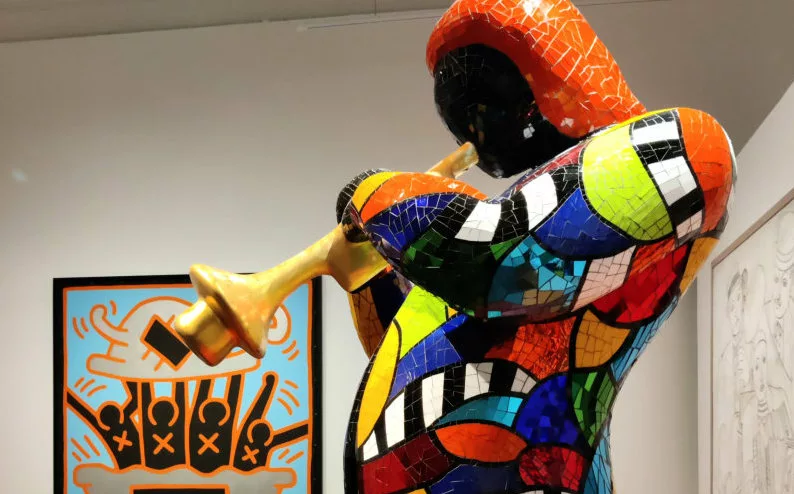 Une sculpture recouverte de mosaïque miroir colorées représentant un homme en veste en train de jouer de la trompette (et dans le fond un tableau)