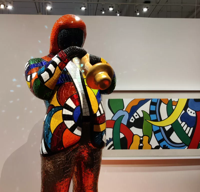 Une sculpture recouverte de mosaïque miroir colorées représentant un homme en veste en train de jouer de la trompette (et dans le fond un tableau)