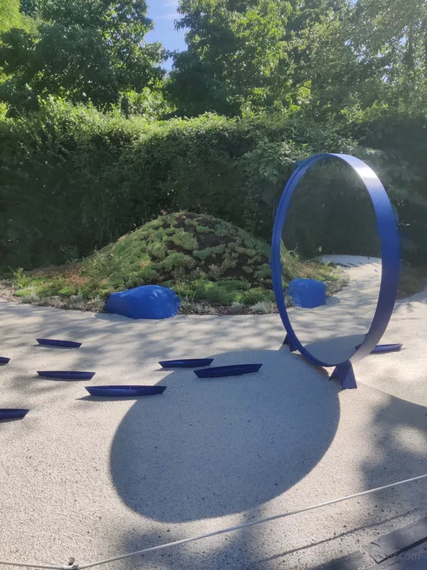 Un parterre en sable sur lequel sont posés de petites barques bleues et un miroir rond bleu. Dans le fond, un rocher bleu précède un talus végétalisé