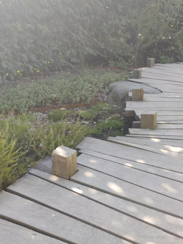 Un chemin fait de planches en bois de tailles et de formes différentes. Certaines sont découpées au plus juste pour contourner un cailloux