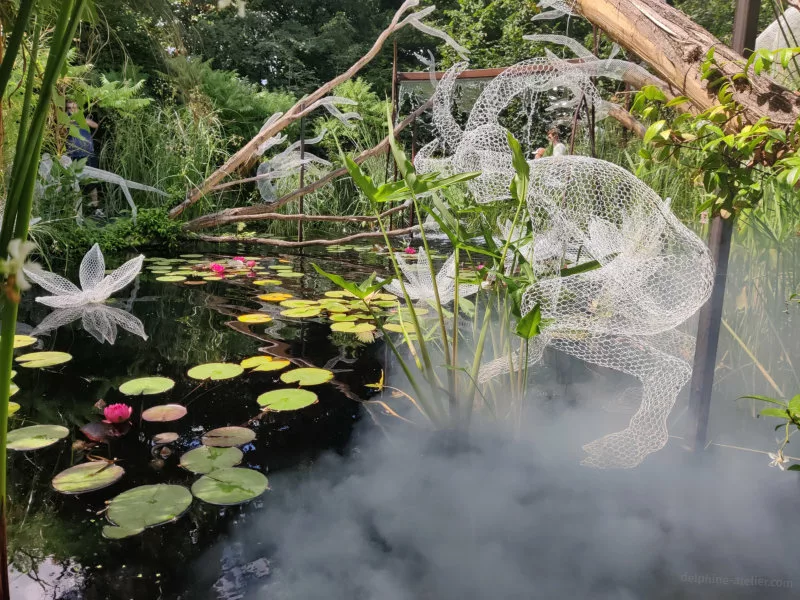 Une créature faite de grillage blanc se penche au-dessus d'une mare avec des nénuphars dans un peu de fumée blanche