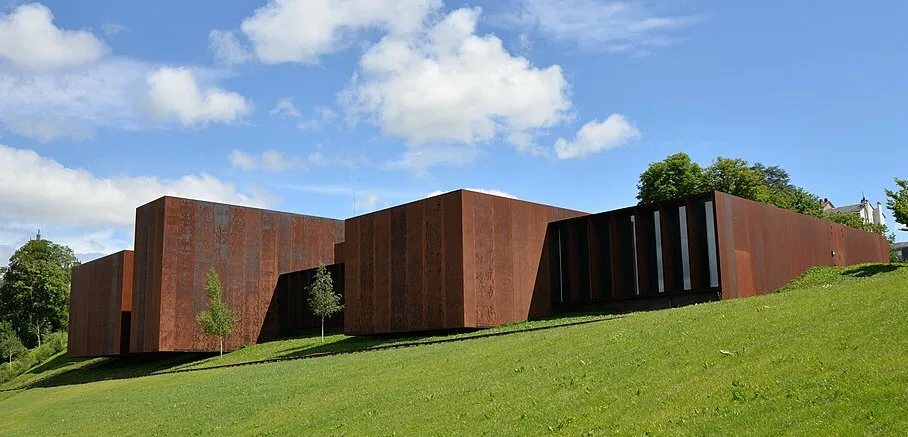 Un bâtiment fait de parallélépipèdes brun-rouille qui semble posé sur une butte d'herbe