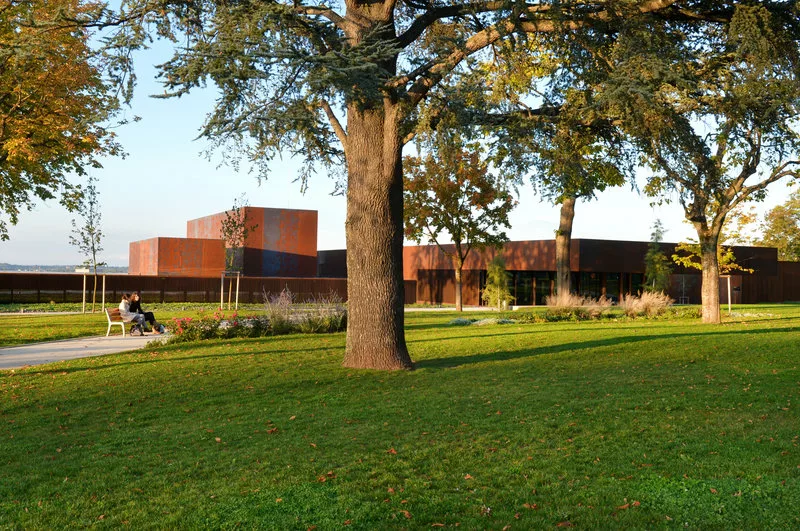 Dans un parc arboré où deux personnes discutent sur un banc, des blocs d'un bâtiment cuivré donnent l'impression d'un bâtiment quasi plat d'un niveau seulement