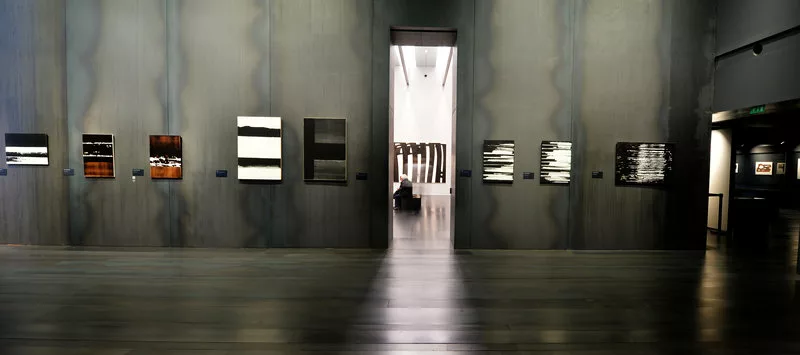 Une salle d'exposition aux murs et au sol noir donne accès à une salle aux murs blancs. On voit que les panneaux de murs noirs ne sont pas tout à fait unis mais qu'il y a des traces comme des réactions du matériaux.