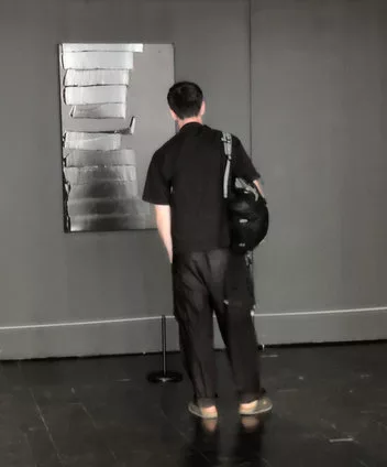 Un homme brun, habillé de noir avec un sac noir et des chaussures noires (gris foncé ?) regarde un tableau entièrement noir (même si les reflets de la lumière donnent beaucoup de blanc) accroché sur un mur noir et avec un sol... noir