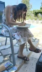 Une jeune fille en train peindre à l'aquarelle (on voit la boîte par terre) dans un carnet au bord d'une piscine