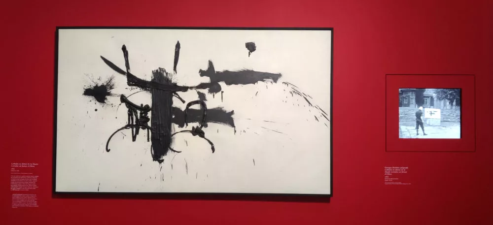 Un tableau accroché sur un mur à côté duquel un écran diffuse des images du même tableau inachevé et l'artiste devant