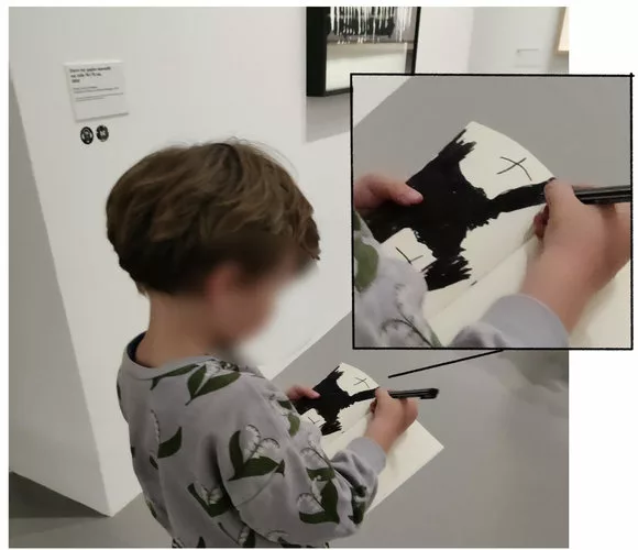 Un enfant (visage flouté) marche dans un musée en train de dessiner debout. Un zoom permet de voir qu'il colore une page entière en noire à l’exception de deux zones avec une croix