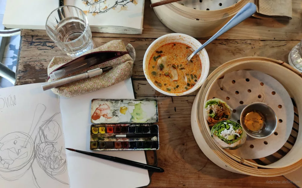 Une table de repas (soupe, rouleaux de printemps) et de dessin en cours (carnets, aquarelle, etc.)