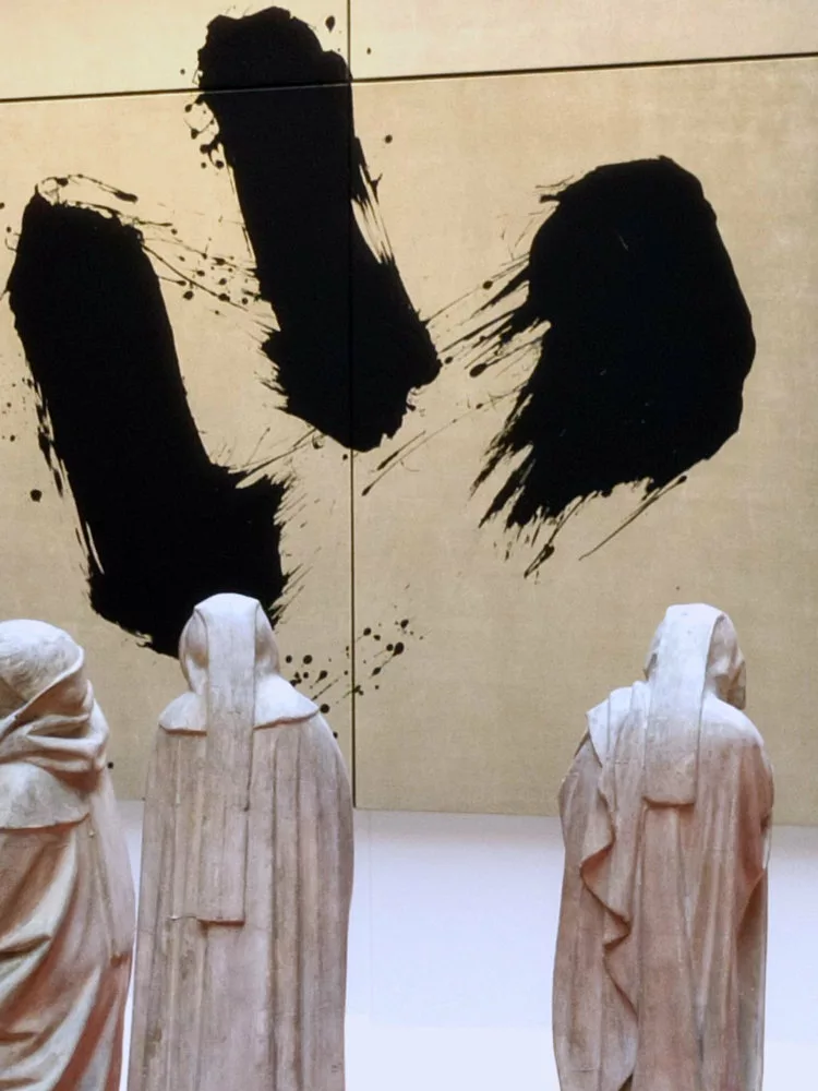 À l'arrière plan, une toile à fond doré et trois traces dynamiques noires ; au premier plan, des statuettes de moines qui nous tournent le dos