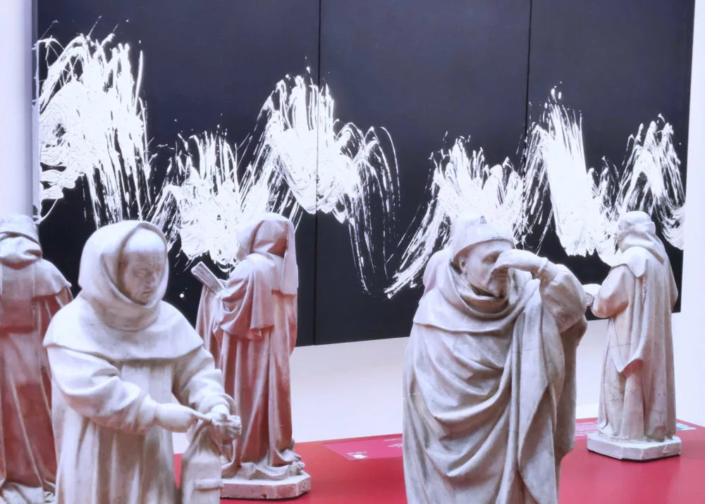 À l'arrière plan, une toile bleue avec des traces ondulantes dynamiques blanches ; au premier plan, des statuettes de moines dont un qui se bouche le nez