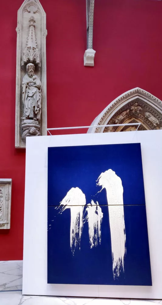 À l'arrière plan, un élément architectural sculpté tout en hauteur ; au premier plan, une paroi éphémère sur laquelle est accrochée une toile bleue avec trois traces verticales blanches