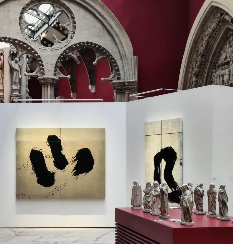 À l'arrière plan, des éléments architecturaux ; au second plan, des parois éphémères sur lesquelles sont accrochées des toiles à fond or et traces noires ; au premier plan en biais, un socle présentant toute une série de statuettes de moines à capuche