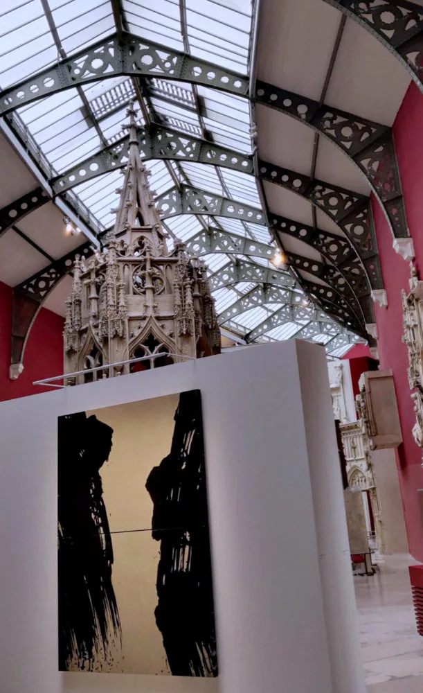 En arrière plan, une verrière posée sur une structure métallique en pointe ; au second plan, un élément architectural, au premier plan une paroi éphémère sur laquelle est accroché une toile de Fabienne Verdier à fond or et traces noires