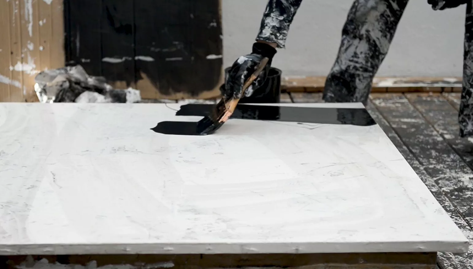 Une toile blanche est posée au sol. On voit une artiste en train de tracer une ligne noir brillant à l'aide d'un pinceau très large