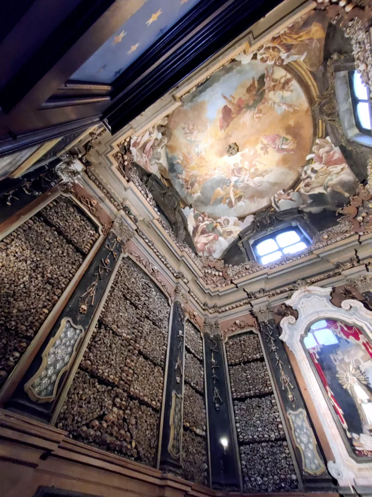 Photo d'une chapelle en conte-plongée où on voit un plafond peint et des murs recouverts d'ossements agencés en décoration