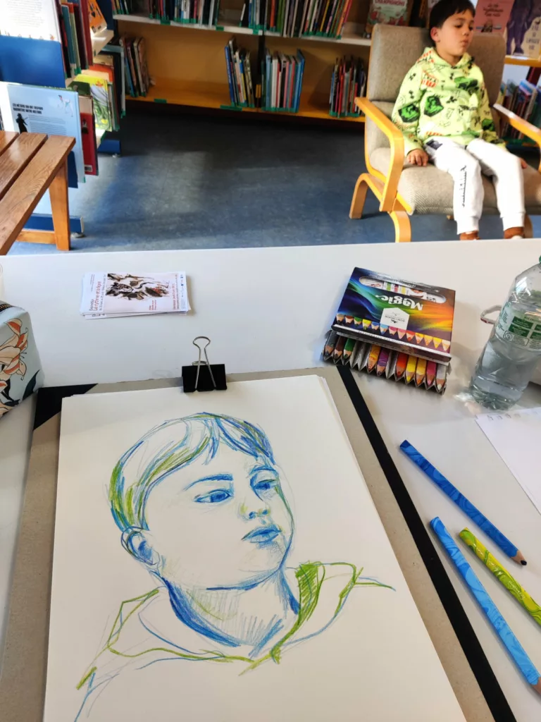 Une table de dessinatrice avec le portrait d'un enfant fini et des crayons de couleurs ; au deuxième plan, l'enfant qui pose (en bougeant :D )