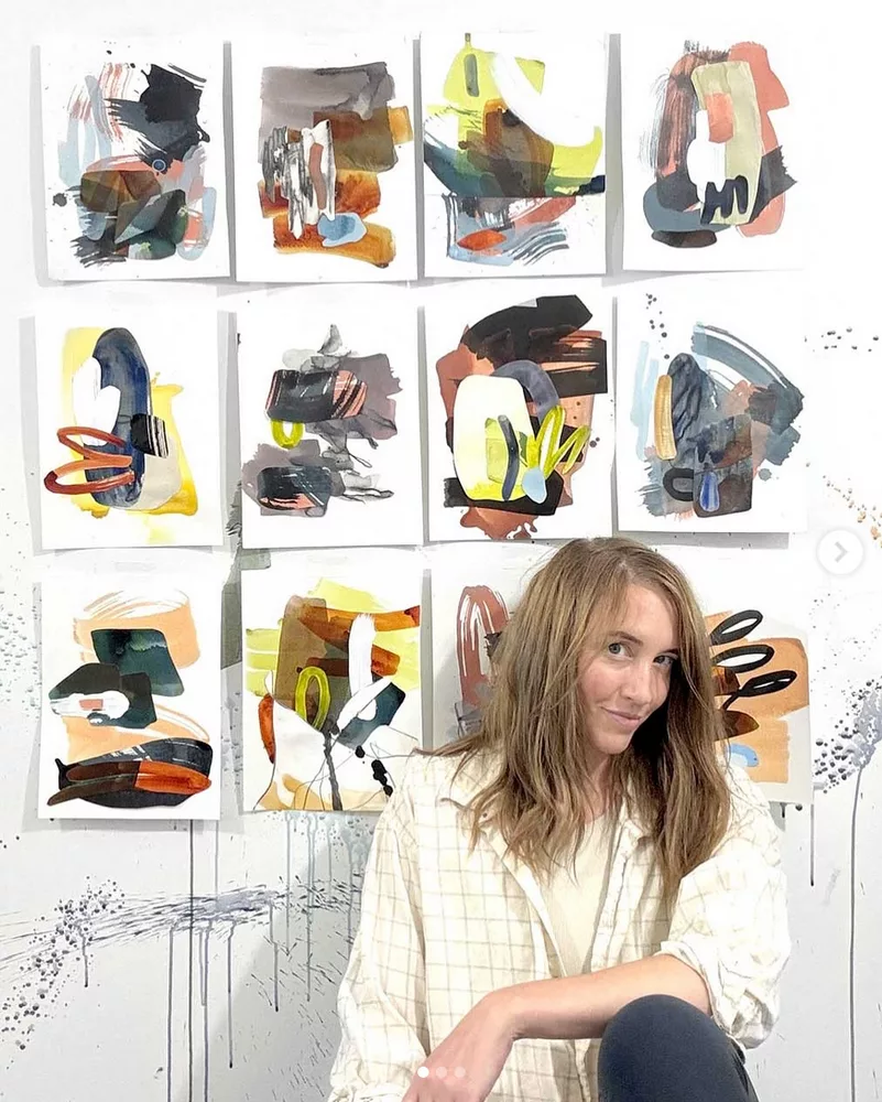 Une femme souriante pose devant douze feuilles sur lesquelles il y a une composition de couleurs et de formes